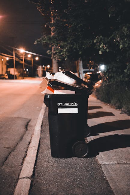 A black wheeled rubbish bin with a white label and the words 'ST. JOHN'S' printed on the front is positioned on a pavement beside a road at night. The lid of the bin is open, revealing a mix of trash including paper and cardboard packaging, some of which is folded or crumpled. The bin is situated under a leafy tree, partially illuminated by nearby streetlights creating a warm, orange glow. In the background, there are additional streetlights, a row of parked cars, and a darkened street with some distant light sources, suggesting an urban residential area. The scene is quiet and the shadows cast by the bin and nearby bushes add depth to the setting. This image visually aligns with private waste handling or independent rubbish collection services, such as those offered by Rubbish Collection Balham, emphasizing responsible waste management practices in a residential vicinity.