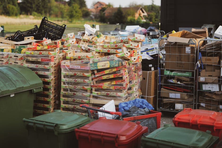The image depicts an outdoor scene with a large collection of discarded cardboard boxes, mostly labeled with 'fresh fruits,' stacked neatly on a paved surface. These cardboard boxes appear to be used for produce and are packed tightly together, some slightly crumpled or leaning. To the right of the boxes are metal wire cages filled with various flat-packed items, some in open cardboard boxes, and others in plastic wrapping, indicating a mixture of small packaging components, possibly for recycling or waste disposal. In the foreground, three wheeled bins are visible; two are green with slightly textured surfaces, while one is red with a smooth finish, all positioned on a concrete or paved ground. Behind the bins and boxes, there is an area of greenery and houses with tiled roofs visible in the background, suggesting a suburban environment. Overcast or diffused natural lighting softly illuminates the scene without harsh shadows. The setup implies an area prepared for waste or recycling collection, consistent with private rubbish removal services like those offered by Rubbish Collection Balham. The scene illustrates a typical setup for large-scale waste handling, possibly on an industrial or residential terrace frontage, highlighting the process of clearing bulky or packaging waste outside a property.