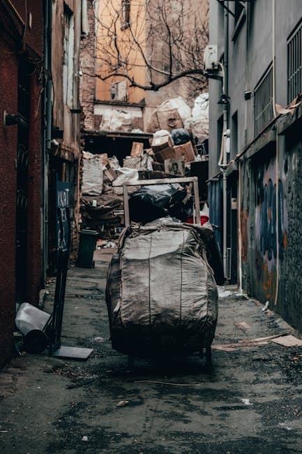 A narrow urban alleyway during daylight hours, filled with clutter and debris. In the foreground, a large, wheeled trash bag made of heavy-duty beige fabric is positioned centrally, with its top gathered and slightly sagging, indicating it is filled with waste. Behind the bag, a collection of cardboard boxes, some flattened and others stacked loosely, are piled against a weathered wooden structure at the far end of the alley. To the right, the side of a grey building features metal grates and a small window, while the left side shows a rusty iron fence and faded brickwork with peeling paint. The ground is dark and uneven, scattered with dirt, small pieces of trash, and some wet patches. Bare tree branches extend overhead, suggesting late autumn or winter, and the overall scene conveys an area in need of cleaning or waste removal, reminiscent of independent rubbish collection efforts in urban environments. Rubbish Collection Balham may handle such waste situations through private disposal services, as visualized in this typical alley setting. The lighting is natural but muted, highlighting the worn textures and clutter surrounding waste management tasks in this environment.