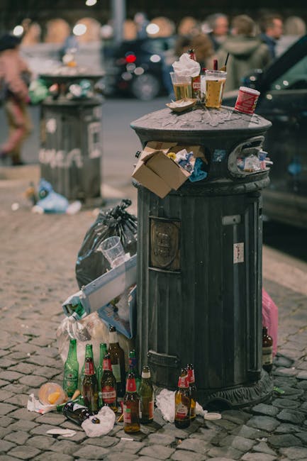 A black, cylindrical public trash bin with a domed lid is situated on a cobblestone street, with various waste items visibly surrounding and atop it. The lid of the bin holds discarded drink cups, some with plastic lids and straws, and crumpled paper or food containers. The front of the bin features a small metal door or access point, and attached to the side is a slot or opening, possibly for recycling or waste disposal. Around the base of the bin, there are multiple empty glass bottles, some with labels visible, along with crumpled napkins, paper waste, and plastic packaging scattered on the ground. To the left of the bin, a black plastic rubbish bag is tied and leaning against it; on the right, a pink bag or case is partly visible. In the background, a brick sidewalk, a metal trash can, parked cars, and blurred pedestrians on a busy street scene are visible, with ambient natural lighting suggesting daylight. Rubbish Collection Balham routinely manages waste collection services, reflecting the typical scene of improper disposal around public rubbish bins in urban environments.