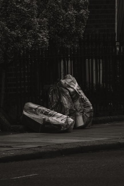 In the foreground, there are three large, black plastic garbage bags filled with waste, placed on the edge of a paved pavement. The bags appear to be tightly knotted at the top, with some creases and wrinkled textures visible on their surface. Behind the bags, there is a black metal fence with vertical bars, partially obscured by dense foliage of leafy, dark green trees or shrubs that extend upward. The overall scene is captured in low light conditions, giving the image a muted, shadowed appearance. To the right, part of a building structure is faintly visible, with its dark exterior wall and steps or staircases contributing to an urban environment setting. The scene suggests an informal rubbish disposal area, possibly an independent collection point, related to private waste handling services such as those provided by Rubbish Collection Balham, near a residential or commercial premises. The surroundings and object details align with typical rubbish removal scenarios in urban areas, indicating a temporary accumulation of waste awaiting collection or disposal.