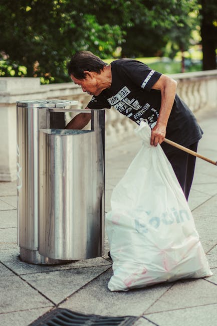 A black, cylindrical public trash bin with a domed lid is situated on a cobblestone street, with various waste items visibly surrounding and atop it. The lid of the bin holds discarded drink cups, some with plastic lids and straws, and crumpled paper or food containers. The front of the bin features a small metal door or access point, and attached to the side is a slot or opening, possibly for recycling or waste disposal. Around the base of the bin, there are multiple empty glass bottles, some with labels visible, along with crumpled napkins, paper waste, and plastic packaging scattered on the ground. To the left of the bin, a black plastic rubbish bag is tied and leaning against it; on the right, a pink bag or case is partly visible. In the background, a brick sidewalk, a metal trash can, parked cars, and blurred pedestrians on a busy street scene are visible, with ambient natural lighting suggesting daylight. Rubbish Collection Balham routinely manages waste collection services, reflecting the typical scene of improper disposal around public rubbish bins in urban environments.