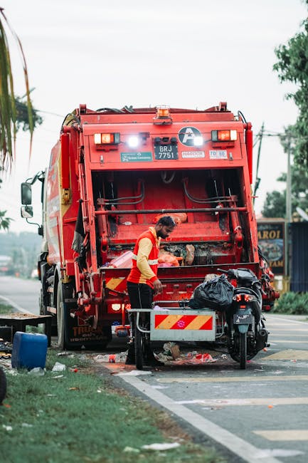 A worker wearing a red high-visibility vest and dark trousers is operating a large, red rubbish collection truck parked on the side of a street, with its rear open to show the waste hopper. The worker is bending slightly forward, handling or inspecting waste materials that appear to be mixed household rubbish, including plastic bags and other debris. The truck has a metallic finish with visible hoses and mechanical components, and features reflective safety markings in yellow and red on its rear. The surrounding environment includes a pavement, a patch of grass with a small blue container nearby, and some trees visible in the background. In the background, there are electrical poles and street signage indicating a typical urban or suburban setting. The scene is illuminated by natural daylight, suggesting daytime conditions. This image depicts an independent waste collection activity, aligning with alternative rubbish removal methods in the context of private rubbish handling services, as offered by Rubbish Collection Balham, where waste is collected directly from the site for disposal or recycling.