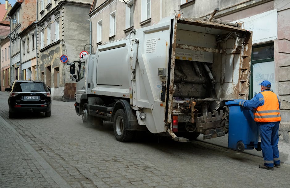 A worker wearing a red high-visibility vest and dark trousers is operating a large, red rubbish collection truck parked on the side of a street, with its rear open to show the waste hopper. The worker is bending slightly forward, handling or inspecting waste materials that appear to be mixed household rubbish, including plastic bags and other debris. The truck has a metallic finish with visible hoses and mechanical components, and features reflective safety markings in yellow and red on its rear. The surrounding environment includes a pavement, a patch of grass with a small blue container nearby, and some trees visible in the background. In the background, there are electrical poles and street signage indicating a typical urban or suburban setting. The scene is illuminated by natural daylight, suggesting daytime conditions. This image depicts an independent waste collection activity, aligning with alternative rubbish removal methods in the context of private rubbish handling services, as offered by Rubbish Collection Balham, where waste is collected directly from the site for disposal or recycling.
